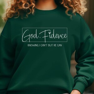 以下が含まれることがあります： 白い書体で「God. Fidence」と書かれた濃い緑色のスウェットシャツ。テキストは白い長方形で囲まれています。長方形の下には「Knowing I can't but He can.」というテキストがあります。