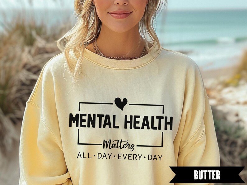 Peut inclure: Un sweat-shirt jaune clair avec l'inscription noire "MENTAL HEALTH Matters ALL DAY EVERY DAY". Un c&oelig;ur noir est au-dessus du mot "HEALTH". Le mot "BUTTER" est dans une banni&egrave;re noire en bas.