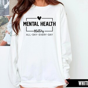 Peut inclure: Sweat-shirt blanc avec l'inscription noire "MENTAL HEALTH Matters ALL DAY EVERY DAY", un petit c&oelig;ur noir au-dessus de "MENTAL". Le sweat-shirt est &agrave; manches longues et col rond.