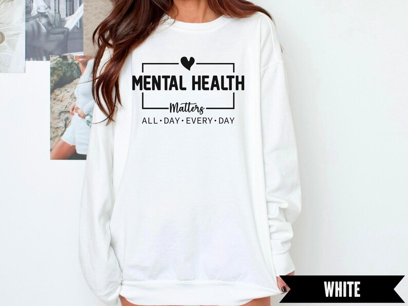 Peut inclure: Sweat-shirt blanc avec l'inscription noire "MENTAL HEALTH Matters ALL DAY EVERY DAY", un petit c&oelig;ur noir au-dessus de "MENTAL". Le sweat-shirt est &agrave; manches longues et col rond.