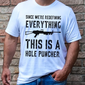 May include: White t-shirt with a black graphic of a machine gun and the text "Since we're redefining everything, this is a hole puncher."