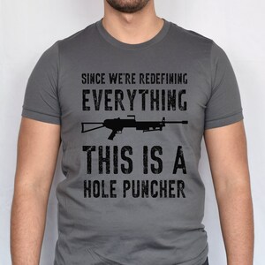 May include: A dark gray t-shirt with a black graphic print of a rifle and the text "Since we're redefining everything, this is a hole puncher".