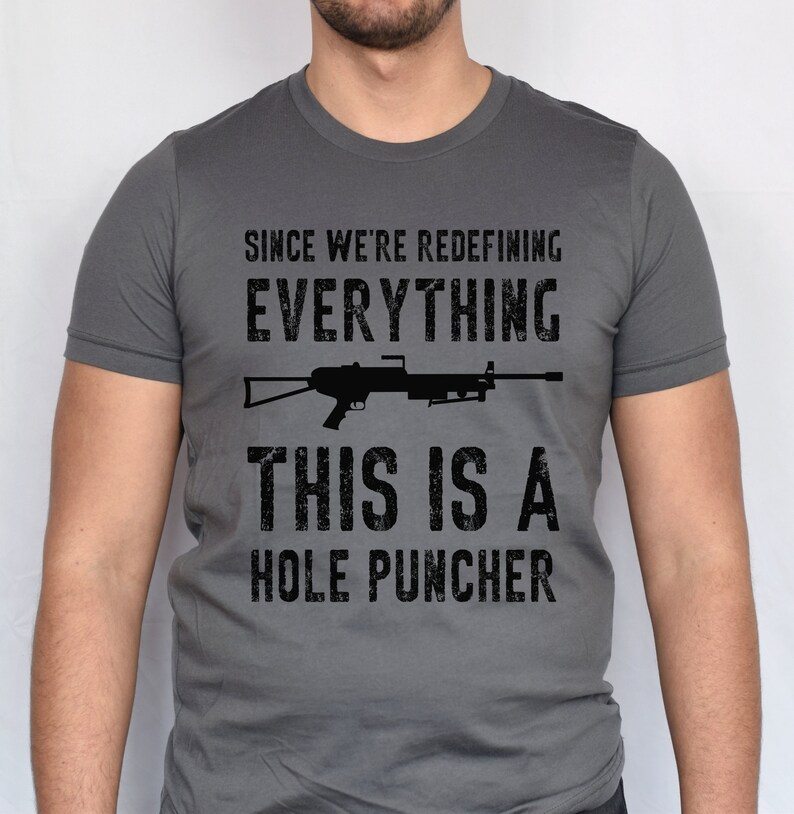 May include: A dark gray t-shirt with a black graphic print of a rifle and the text "Since we're redefining everything, this is a hole puncher".