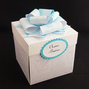 May include: A white gift box with a blue and white bow on top. The box has a blue oval sticker with the text "On your Baptism" on it.