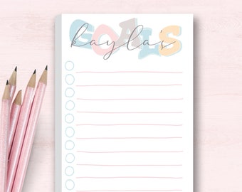 Puede incluir: Un bloc de notas blanco con la palabra "GOALS" en colores pastel y el nombre "Kayla" en cursiva. El bloc de notas tiene líneas horizontales rosas y círculos azul claro en el lado izquierdo. Lápices rosas a la izquierda.