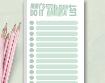 Puede incluir: Un bloc de notas blanco con el texto "Abby's Do It Today List" en verde claro. El bloc de notas tiene una serie de círculos y líneas verdes claros para una lista de tareas. Lápices rosas están a la izquierda del bloc de notas.