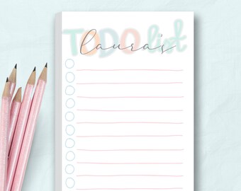 Puede incluir: Un bloc de notas blanco con las palabras "TO DO list" en colores pastel y el nombre "Laura's" en cursiva. Lápices rosas están colocados en el lado izquierdo del bloc de notas. El bloc de notas tiene líneas horizontales rosas y círculos azules.