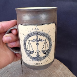 Op de afbeelding: Bruine keramische mok met een Weegschaal sterrenbeeld ontwerp. Het ontwerp toont een weegschaalsymbool, omgeven door een cirkelvormig frame met stralende lijnen en het woord "LIBRA". De mok heeft een handvat en een crèmekleurig interieur.