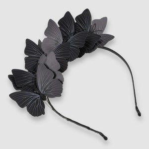 Puede incluir: Una diadema negra adornada con múltiples adornos de mariposas. Las mariposas son de varios tonos de negro y gris, con delicados detalles de venas blancas en sus alas. La diadema es de un color oscuro y elegante.