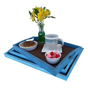 May include: Two blue wooden trays with a brown woven mat inside. The top tray has a donut, a white cup with pink and red candy hearts, a white mug with blue and white design, and a white saucer. A vase with yellow flowers is in the background.