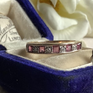 May include: A gold ring set with pink gemstones and small diamonds, presented in an open, vintage blue velvet ring box. The ring box has a cream-coloured interior. A white flower is visible in the background.