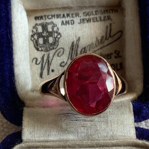 May include: A gold ring featuring a large, oval-cut, deep red gemstone set in a blue velvet-lined jewelry box. The interior of the box lid displays the printed text: WATCHMAKER, GOLDSMITH AND JEWELLER, W. Mansell, Silver St, Lincoln.