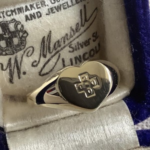 Peut inclure: Une bague en or en forme de cœur avec un motif de croix sur le devant. La bague est dans un écrin à bague en velours bleu avec une étiquette blanche qui indique "Watchmaker, Goldsmith and Jeweller W. Mansell Silver St. Lincoln."