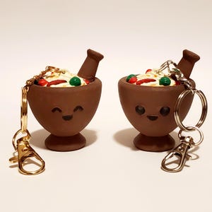 May include: Two brown mortar and pestle keychains with smiling faces, filled with white, red, and green food. Each keychain has a gold or silver clasp and ring.