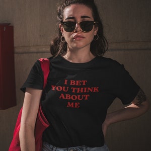 Op de afbeelding: Een zwart T-shirt met de tekst "I bet you think about me" in rood.