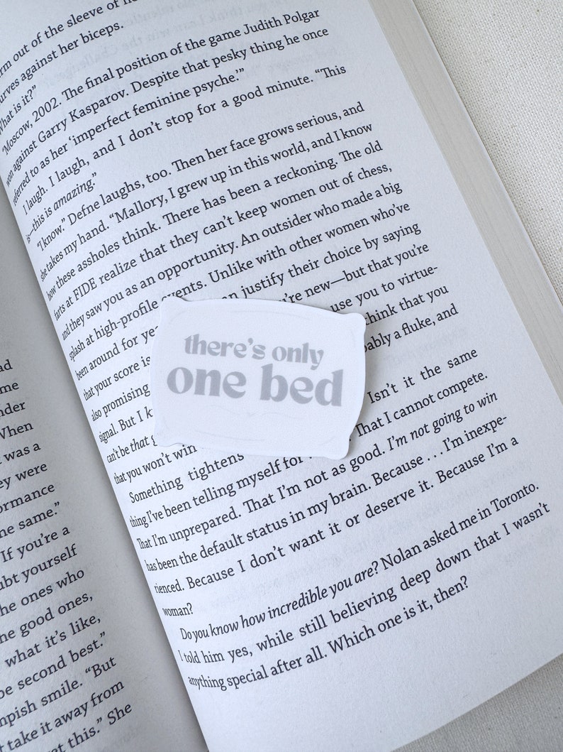 Op de afbeelding: Een close-up van een boekpagina met de tekst "there's only one bed" gedrukt op een witte sticker. De tekst is in een vet, sans-serif lettertype. De sticker is geplaatst over een paragraaf tekst over de ervaring van een vrouw in de schaakwereld.