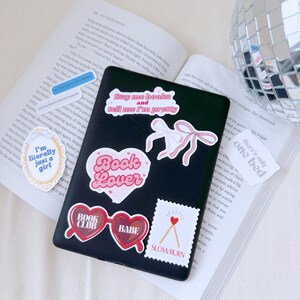 Op de afbeelding: Een zwarte laptop met verschillende stickers. De stickers zijn roze, rood en wit en bevatten zinnen als "Koop me boeken en vertel me dat ik mooi ben", "Boekenliefhebber", "Boekenclub", "Babe" en "Slowburn".