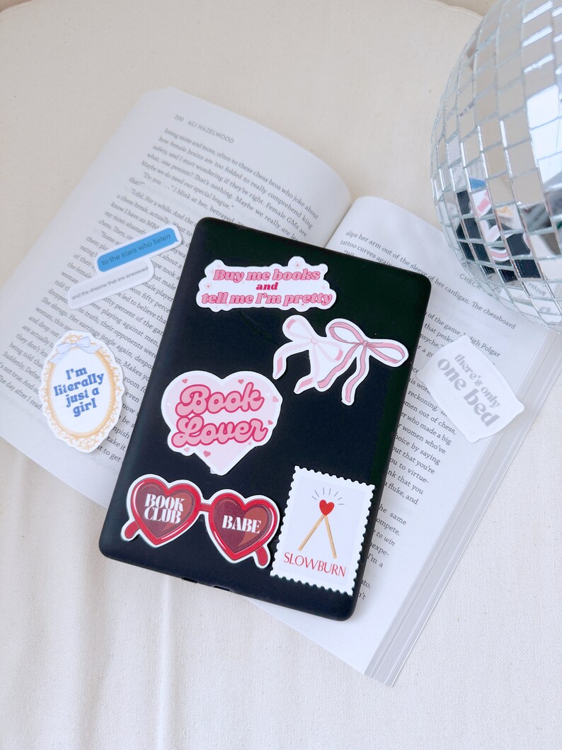Op de afbeelding: Een zwarte laptop met verschillende stickers. De stickers zijn roze, rood en wit en bevatten zinnen als "Koop me boeken en vertel me dat ik mooi ben", "Boekenliefhebber", "Boekenclub", "Babe" en "Slowburn".