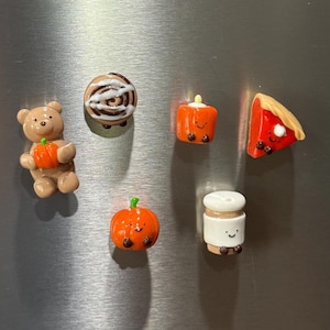 Puede incluir: Un conjunto de seis imanes en miniatura que incluye un oso marrón con una calabaza naranja, un rollo de canela, un cuadrado naranja con cara, una porción de tarta roja, una calabaza naranja con cara y una taza de café blanca con cara.
