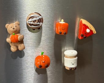 Puede incluir: Un conjunto de seis imanes en miniatura que incluye un oso marrón con una calabaza naranja, un rollo de canela, un cuadrado naranja con cara, una porción de tarta roja, una calabaza naranja con cara y una taza de café blanca con cara.