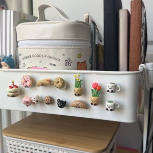 Puede incluir: Un estante de carrito de almacenamiento de metal blanco con un estuche color crema con cremallera y el texto Bobbie Goods Ohuhu, varios imanes pequeños de comida y plantas con caras sonrientes y varios cuadernos verticales en la parte trasera.