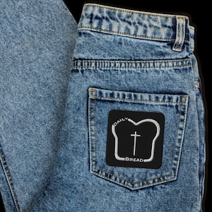 May include: A pair of blue denim jeans with a black patch on the back pocket. The patch features a white outline of a slice of bread with a cross inside and the text "DAILY BREAD".