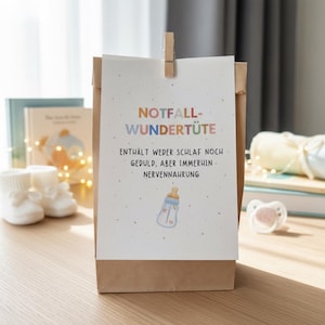 May include: A brown paper gift bag with a white card attached by a wooden clip. The card has text in German and a baby bottle illustration. Baby booties and a pacifier are visible in the background.