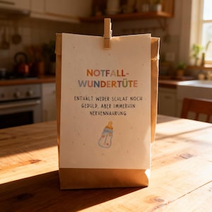 May include: A brown paper bag with a cream-colored card attached by a wooden clip. The card has the words "NOTFALL-WUNDER-T&Uuml;TE" in orange and blue, with German text below and a baby bottle illustration.