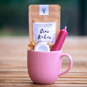 Könnte beinhalten: Eine rosa Keramiktasse gefüllt mit einem braunen Papierbeutel mit heißer Schokolade, einer weißen Kerze und getrockneten Blumen. Der Beutel hat ein weißes Etikett mit der Aufschrift "Heiße Schokolade Ciao Kakao Wir werden dich vermissen!"