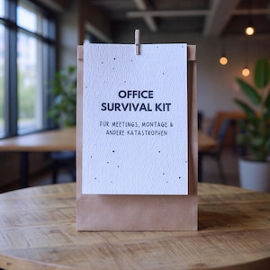 May include: A brown paper bag with a white card clipped to the top. The card reads "OFFICE SURVIVAL KIT" in black text, with additional German text below. The bag sits on a wooden table.
