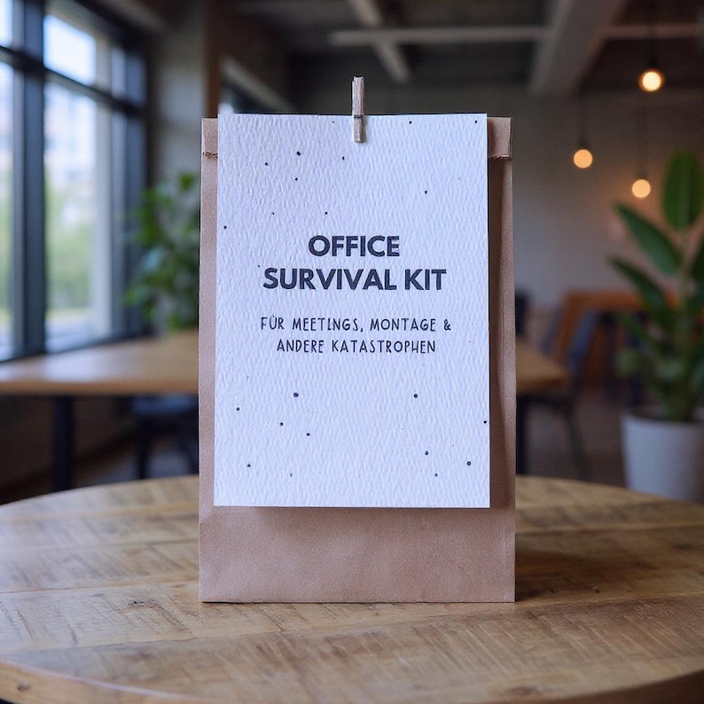 May include: A brown paper bag with a white card clipped to the top. The card reads "OFFICE SURVIVAL KIT" in black text, with additional German text below. The bag sits on a wooden table.