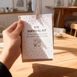 May include: A white card with the text "EHE SURVIVAL KIT" and German text below, held by a wooden clip. The card features a black and white illustration of an elderly couple holding hands. Small black dots are scattered across the card.