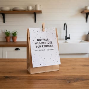 May include: A brown paper bag with a white card clipped to the front. The card has the text "NOTFALL-WUNDER&Uuml;TE F&Uuml;R RENTNER" and "100% FREIZEIT - 0% WECKER" printed on it. The bag is on a wooden surface.