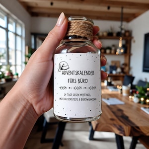Peut inclure: Un bocal en verre transparent avec un goulot entouré de ficelle de jute et une étiquette blanche portant le texte noir ADVENTSKALENDER FÜRS BÜRO, 24 TAGE GEGEN MEETINGS, MOTIVATIONSTIEFS & BÜROWAHNSINN, orné d'un petit bonnet de Père Noël et de points noirs.
