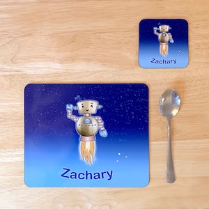 May include: A blue placemat with a starry night sky design features a cartoon robot in a rocket ship. The robot is gold and has a friendly face. The placemat is personalized with the name "Zachary".