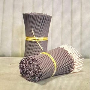 May include: Two bundles of incense sticks. The incense sticks are dark gray and bound with yellow rubber bands. The tips of the incense sticks are white. The bundles are on a light green surface.