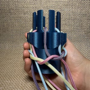 Könnte beinhalten: Eine dunkelblaue Strickliesel aus Kunststoff mit vertikalen Stiften, durch deren Schlitze mehrere bunte Kordeln in Hellblau, Blassgelb, Rosa und Violett gefädelt sind, die an der Unterseite des Werkzeugs herabhängen.