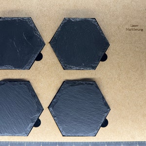 May include: Four black hexagonal slate coasters on a brown surface. The coasters are arranged in a square pattern. The text "Laser Markierung" is visible in the top right corner of the image.