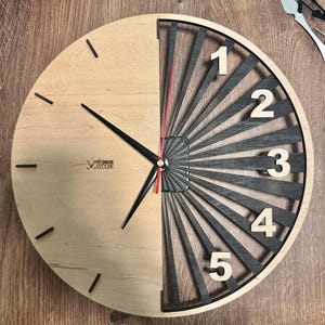 May include: A round wooden wall clock with a unique design. The left side is light wood with black markers, while the right side features a black and gray radial design with white numerals. The clock hands are black with a red second hand. The clock has the text "FOTOWERK STULPE".
