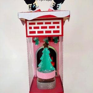 May include: A Christmas-themed decorative box featuring a chimney design with Santa's legs sticking out. Inside, a miniature Christmas tree sits atop a pink and silver base. The box is red and white with brick detailing, and the text "Diseños Arcoiris" is visible.