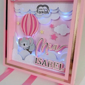 Puede incluir: Caja de sombra rosa con la palabra "Bienvenida" en la parte superior. En el interior, un globo aerostático rosa y blanco transporta un koala sobre las nubes. El nombre "Mia Isabel" se muestra debajo. La caja tiene un borde dorado.