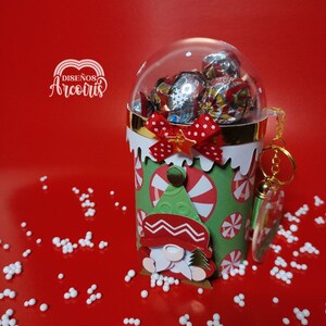May include: A Christmas-themed gift container with a clear dome filled with wrapped candies. The green cylinder features a gnome and candy cane design, accented with a red bow. A small keychain hangs from the side. The background is red.