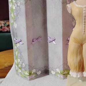 May include: A decorative, three-panel screen with a floral design in shades of lavender, green, and white. The screen features lavender bows and is next to a doll in a yellow outfit. The background includes a framed picture of a ship.