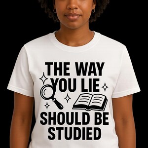 May include: White t-shirt with the text "THE WAY YOU LIE SHOULD BE STUDIED" in black. The design includes a magnifying glass, an open book, and star shapes. The shirt is worn by a person.