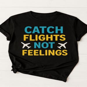 May include: Black t-shirt with the phrase "CATCH FLIGHTS NOT FEELINGS" in teal, yellow, and white. The design includes airplane graphics. The shirt is folded on a white surface.