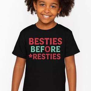 May include: A black t-shirt with the words "BESTIES BEFORE *RESTIES" in red and teal lettering. The shirt is worn by a young person with dark curly hair, smiling at the camera.