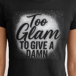 May include: Black t-shirt with the phrase "Too Glam To Give A Damn" in a bold, stylized font. The text is in black and white, with a distressed, speckled background. The shirt is a crew neck.