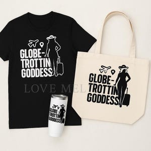 May include: A black t-shirt, a cream tote bag, and a white tumbler featuring the text "GLOBE-TROTTIN GODDESS" with a graphic of a woman with a suitcase, airplane, and location pin. The tumbler is a travel mug.
