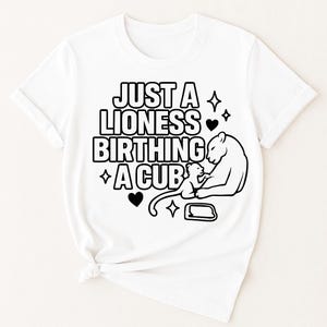 May include: White t-shirt with a black graphic that reads "JUST A LIONESS BIRTHING A CUB" with a lioness and cub illustration. The design includes hearts and star shapes. The shirt has short sleeves and a crew neck.