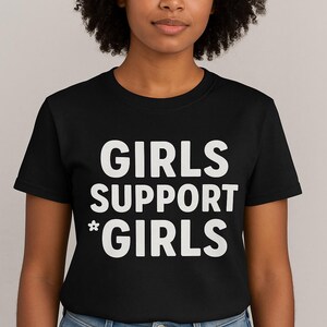 May include: Black t-shirt with the white text "GIRLS SUPPORT *GIRLS". The shirt is a crew neck style and is being worn by a person with dark curly hair and blue jeans.
